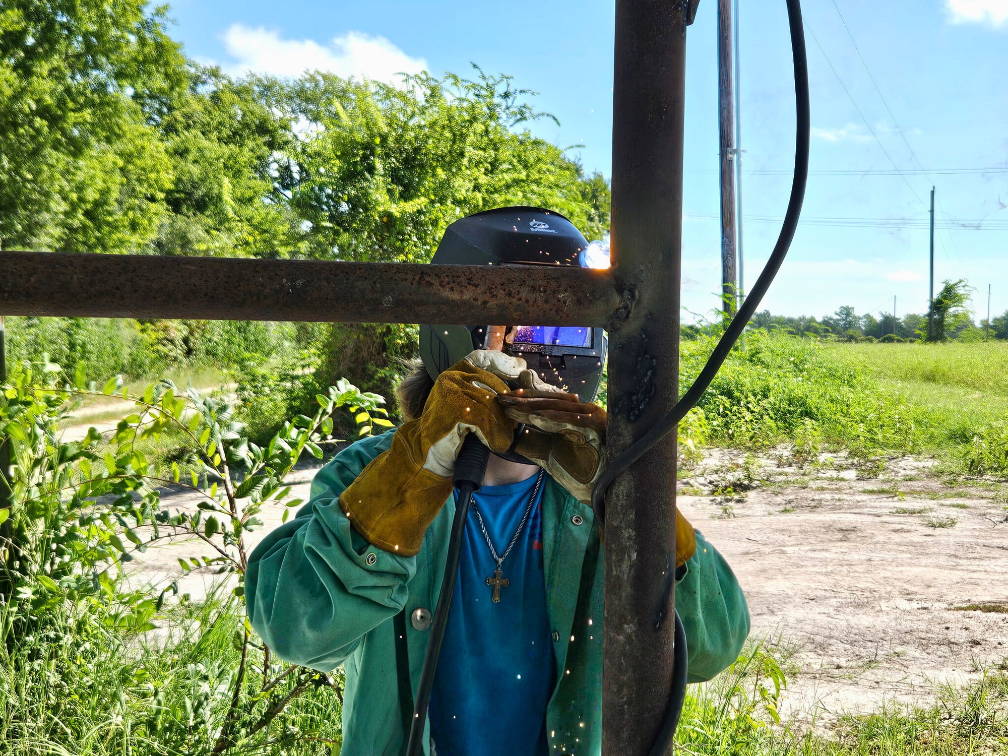 Evan Melvin is wearing protective gear,  working on a metal structure outdoors with greenery and blue sky in the background.